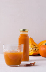 Pumpkin juice pours in a glass glass, next to a bottle of glass on a beige background. In the background is the autumn harvest of pumpkins, copying the space