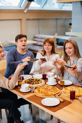 A group of young cheerful friends is sitting in a cafe talking and taking selfies on the phone. Lunch at the pizzeria.