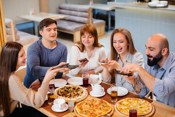 A group of young cheerful friends is sitting in a cafe talking and taking selfies on the phone. Lunch at the pizzeria.