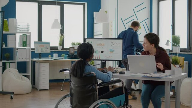 African paralysed invalid immobilized woman in wheelchair and colleague analyzing datas laptop while sitting in start up business office. Colleagues working in background searching financial solutions