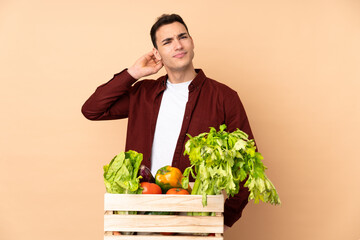 Farmer with freshly picked vegetables in a box isolated on beige background having doubts