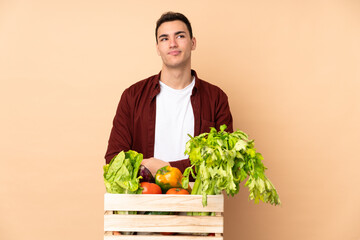 Fototapeta premium Farmer with freshly picked vegetables in a box isolated on beige background making doubts gesture looking side