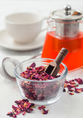 Rose petals craft tea with strainer infuser and clear glass teapot with white ceramic cup on light background.