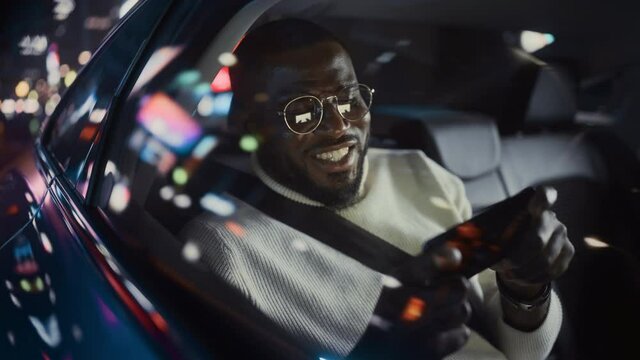 Stylish Black Man in Glasses is Commuting Home in a Backseat of a Taxi at Night. Handsome Male Playing Video Game on Smartphone while in a Car in Urban City Street with Working Neon Signs.