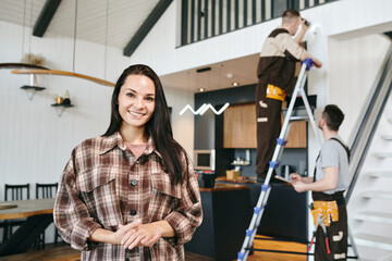 Young cheerful brunette female standing against two repairmen working