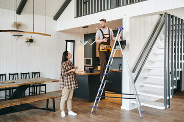 Young man in uniform standing on a ladder in a room and talking to female in casualwear © pressmaster