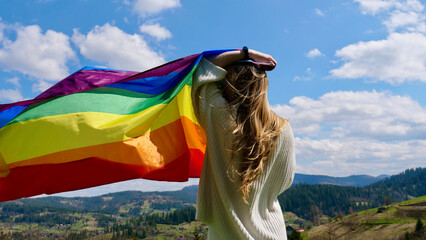 Blonde lesbian woman holding a rainbow LGBT gender identity flag on sky background with clouds on a sunny day