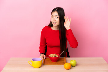 Young Chinese girl  having breakfast in a table listening to something by putting hand on the ear