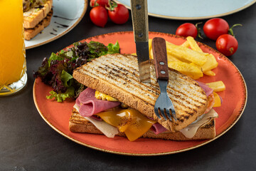 Cheddar cheese, ham and egg toast with orange juice and french fries on a dark stone table.