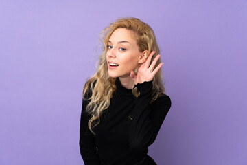 Young blonde woman isolated on purple background listening to something by putting hand on the ear