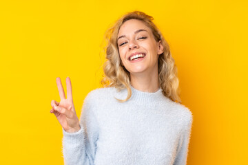 Young blonde woman isolated on yellow background smiling and showing victory sign