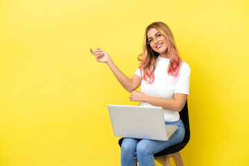 Young woman sitting on a chair with laptop over isolated yellow background extending hands to the side for inviting to come