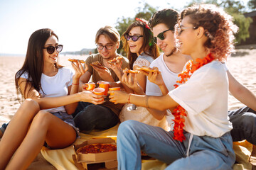 Group of young friends having picnic, eating pizza, toasting with beerus at the beach. Happy friends resting together sitting near the sea. Fast food concept. Beach holiday and summer vacation concept