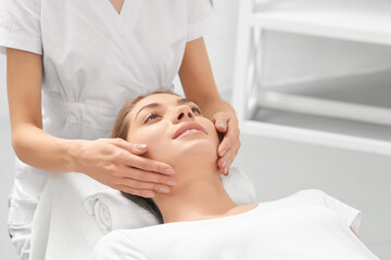 Fototapeta na wymiar Close up portrait of beautician in white uniform doing massage hand for young smiling woman in professional salon. Concept of process special procedure for improvements skin face and body. 