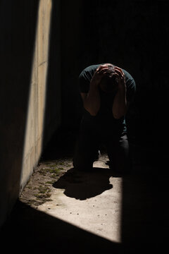 Man kneeling and holding his head with his hands, depressed and desperate in darkness with one sliver of light from above 