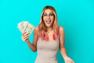 Young woman taking a lot of money over isolated blue background with surprise and shocked facial expression