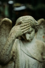 a picture of a statue of an angel, headstone in the cementery, damaged by time, , angel is sad and is touching their head with raised hand