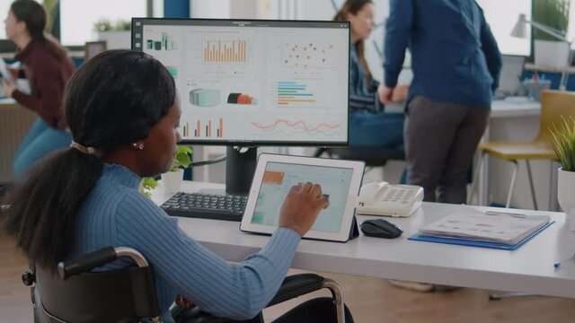Black handicapped woman entrepreneur with locomotor disabilities using computer and tablet in same time working in business start-up office sitting in wheelchair. Busy multitasking african employee