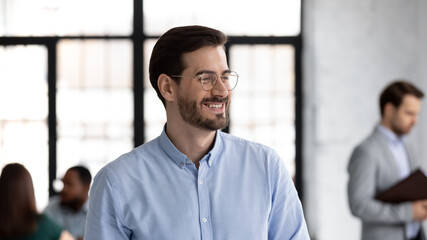 Photo portrait of happy successful business startup leader wearing glasses, looking away, thinking over project, planning future profit and smiling. Multiethnic team working in background. Head shot