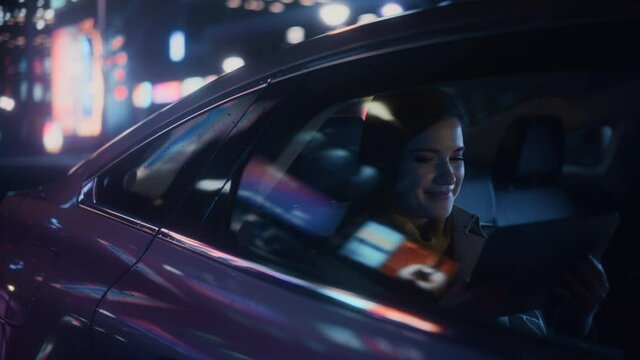 Stylish Female is Commuting Home in a Backseat of a Taxi at Night. Beautiful Woman Passenger Using Tablet Computer and Looking Out of Window while in Car in Urban City Street with Working Neon Signs.