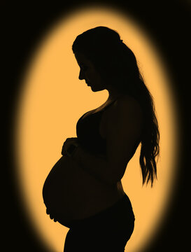 A beautiful pregnant girl with a big belly and long hair stands on a yellow background with an ellipse shadow on the wall. Pregnancy photography.