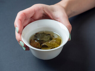 a woman's hand holds a white porcelain bowl with tea. dark background. The opened leaves of large-leaved tea. Chinese tea. tea party
