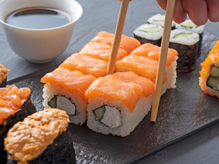 a variety of Japanese rolls and sushi on a textured black plate. Side view. Bamboo chopsticks holding a single roll