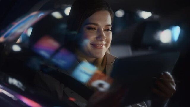 Stylish Female is Commuting Home in a Backseat of a Taxi at Night. Beautiful Woman Passenger Using Tablet Computer and Looking Out of Window while in Car in Urban City Street with Working Neon Signs.