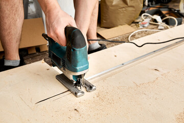 Male hands of construction worker sawing and cutting parts with an electric jigsaw from plywood. The cut process. DIY building of interior decoration with fretsaw. Rental tool. Copy space. Equipment