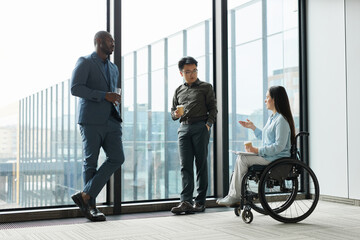 Full length portrait of young businesswoman in wheelchair chatting with male colleagues during...