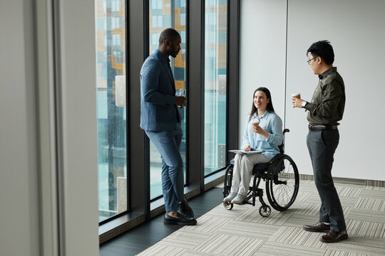 Full length portrait of smiling young businesswoman in wheelchair chatting with male colleagues during coffee break in office