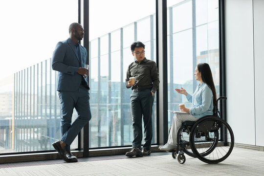Full length portrait of young businesswoman in wheelchair chatting with male colleagues during coffee break in office, copy space