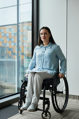 Vertical full length portrait of young businesswoman in wheelchair smiling at camera while posing by window in office