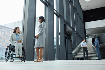 Full length portrait of young businesswoman in wheelchair chatting with female colleague in office lobby, copy space