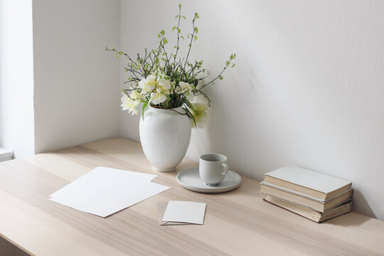 Blank paper sheets, greeting cards on wooden table. Books and cup of coffee. Stationery mockups. Vase with yellow tulips and daffodils. Elegant working space, home office concept. Living room.