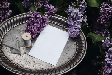 Moody spring wedding stationery mockup scene. Blank greeting card, invitation on vintage silver tray. Purple lilacs flowers and rain drops. Wet silk ribbon. Blooming Syringa bush in the garden. Top.