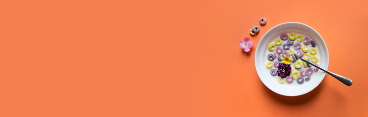 on a peach plain background a beautiful summer American breakfast: multi-colored flakes rings with milk and violet flowers of yellow pink and violet color. there is a spoon in the plate
