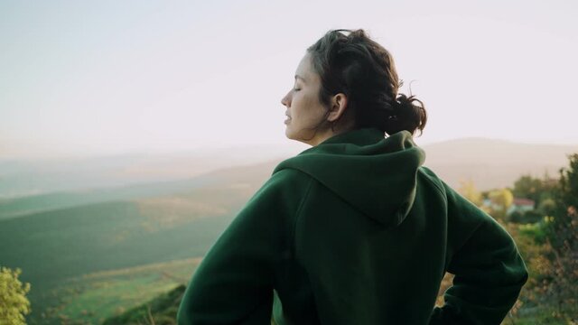 Young woman breathing in fresh air standing on a hill with natural mountains. Green fields and trees in background. She takes a breath relaxing and enjoying nature and beautiful landscape at sunset