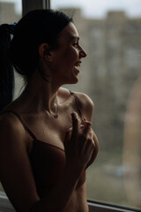 A silhouette of a smiling brunette girl who stands at the window in her lingerie and sprinkles perfume on herself