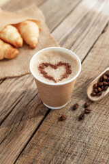 Paper cup of coffee with heart, croissants, beans and spoon at wooden table