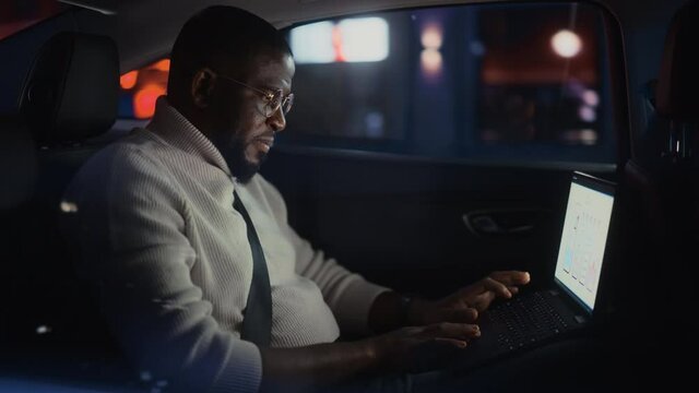 Stylish Black Man in Glasses is Commuting Home in a Backseat of a Taxi at Night. Male Using Laptop Computer and Looking Out of Window while in a Car in Urban City Street with Working Neon Signs.
