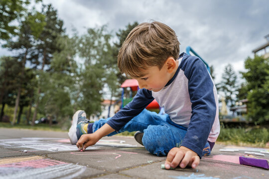 Small caucasian boy four years old sitting on the concrete pavement drawing with chalk in park in day childhood and growing up education concept