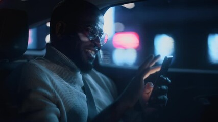 Happy Black Man in Glasses is Commuting Home in a Backseat of Taxi at Night. Handsome Male Using Smartphone and Smiling while in a Car in Urban City Street with Working Neon Signs. Cinematic Footage. - Powered by Adobe