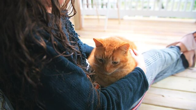 Young woman lying reclining holding in arms on lap sitting cat, petting stroking feline kitty blinking outside at home house balcony porch patio