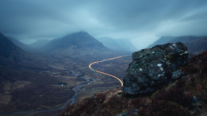 Morning landscape with car lights on the mountain road leading to Glencoe and a big rock in the foreground. A82 road opposite Buachaille Etive Mor before dawn. Scotland © George