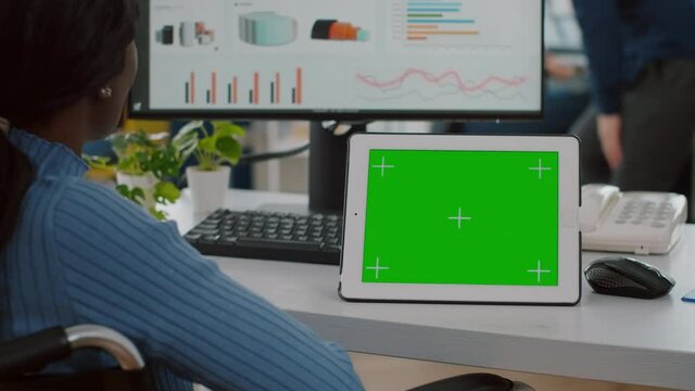 Immobilized african businesswoman in wheelchair looking at digital tablet with green screen chroma key mock up isolated display, comparing graphs from computer working in start up business office