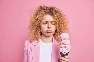 Unhappy curly haired young woman purses lips looks sadly at delicious ice cream feels temptation to eat sweet tasty high calories dessert dressed in formal jacket isolated over pink background