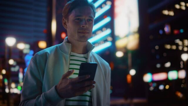 Portrait of Man Using Smartphone Walking Through Night City Street Full of Neon Light. Smiling Stylish Man Using Mobile Phone, Social Media, Online Shopping, Texting on Dating App. Medium Shot