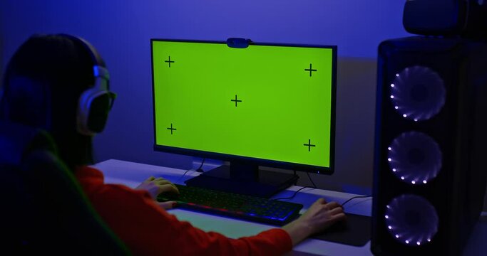 Gamer sits at a large computer monitor with a green screen. Powerful personal computer for computer game lovers, a dark room with illumination. Back view. 4k, ProRes