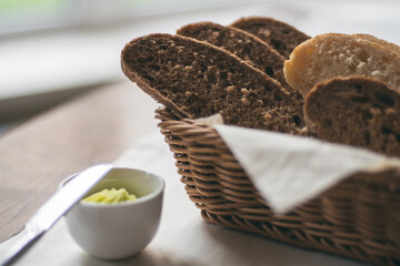 Partially blurred wicker basket with black and white bread and bowl of butter with herbs on white tablecloth near window